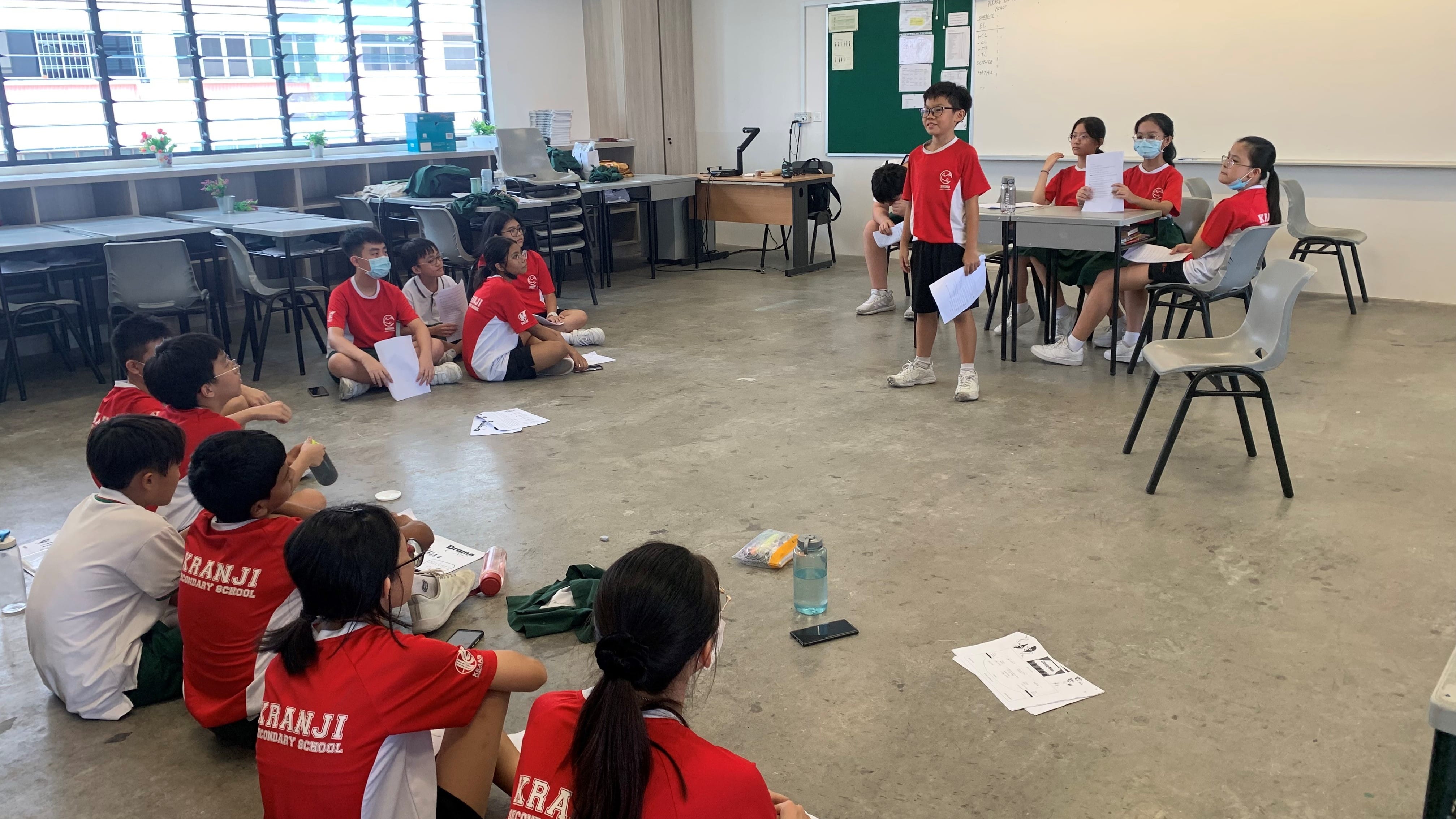 Students in red Kranji Secondary School uniforms sit on a classroom floor watching a peer present papers.